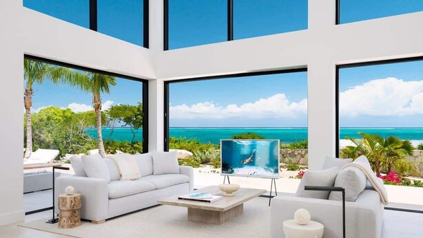Un salon moderne doté d’une grande fenêtre donnant sur une plage, d’un canapé blanc, d’une table basse et d’un écran de télévision diffusant une scène de plage.