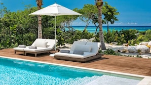 Un espace aménagé au bord de la piscine, avec deux chaises longues et un parasol blanc, entouré d’une végétation tropicale et offrant une vue sur l’océan.