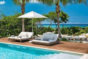 A poolside area with two lounge chairs and a white umbrella, surrounded by tropical vegetation and a view of the ocean.