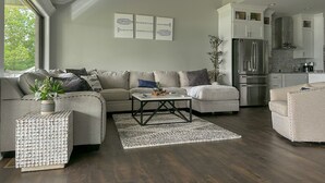 Ein modernes Wohnzimmer mit einer Couch, Couchtisch und Dekoration. Im Hintergrund ist eine Küche mit Edelstahlgeräten zu sehen.