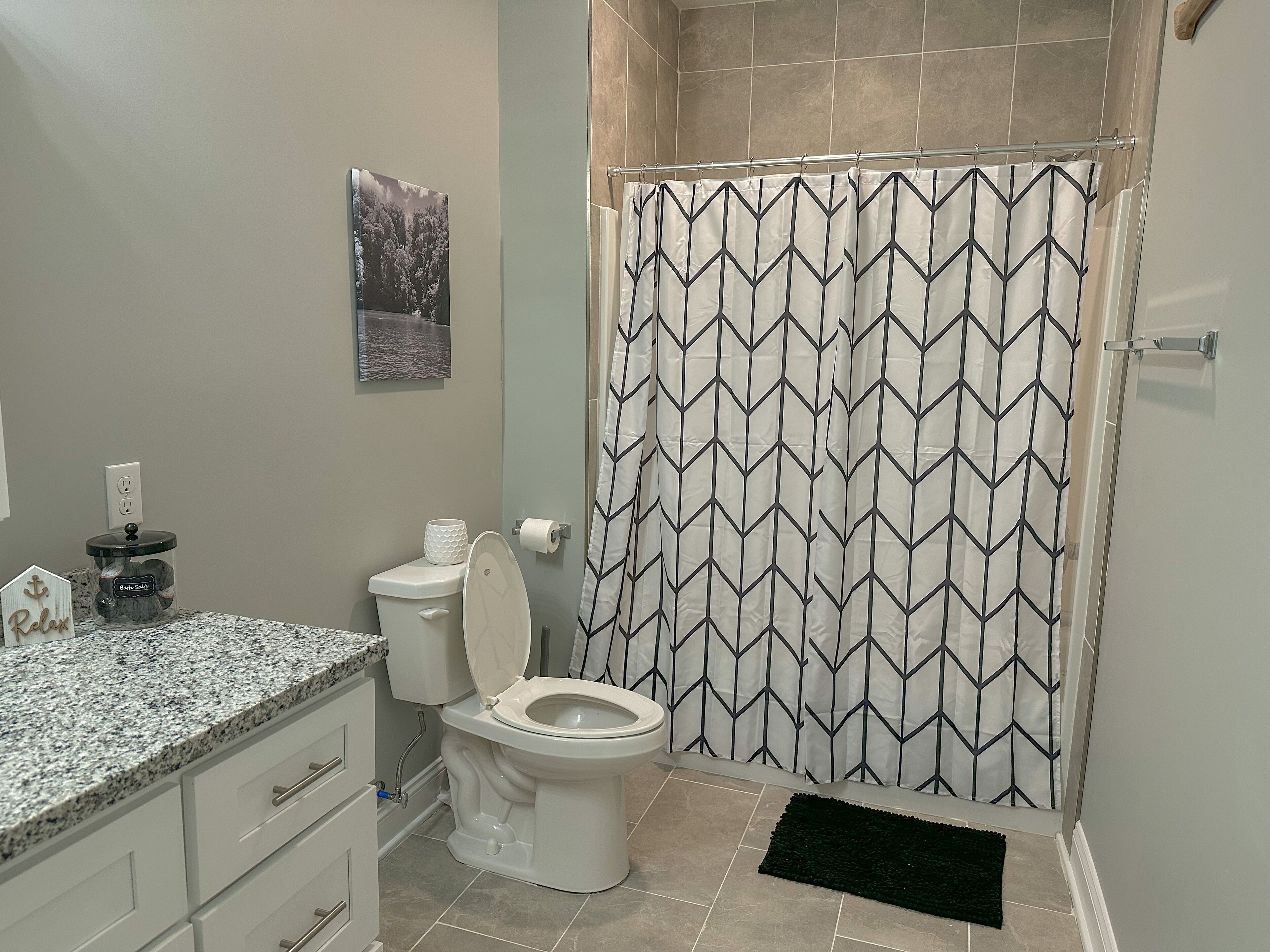 A bathroom with a toilet, a shower curtain with a geometric pattern, a granite countertop, a small trash can, and a framed picture on the wall.
