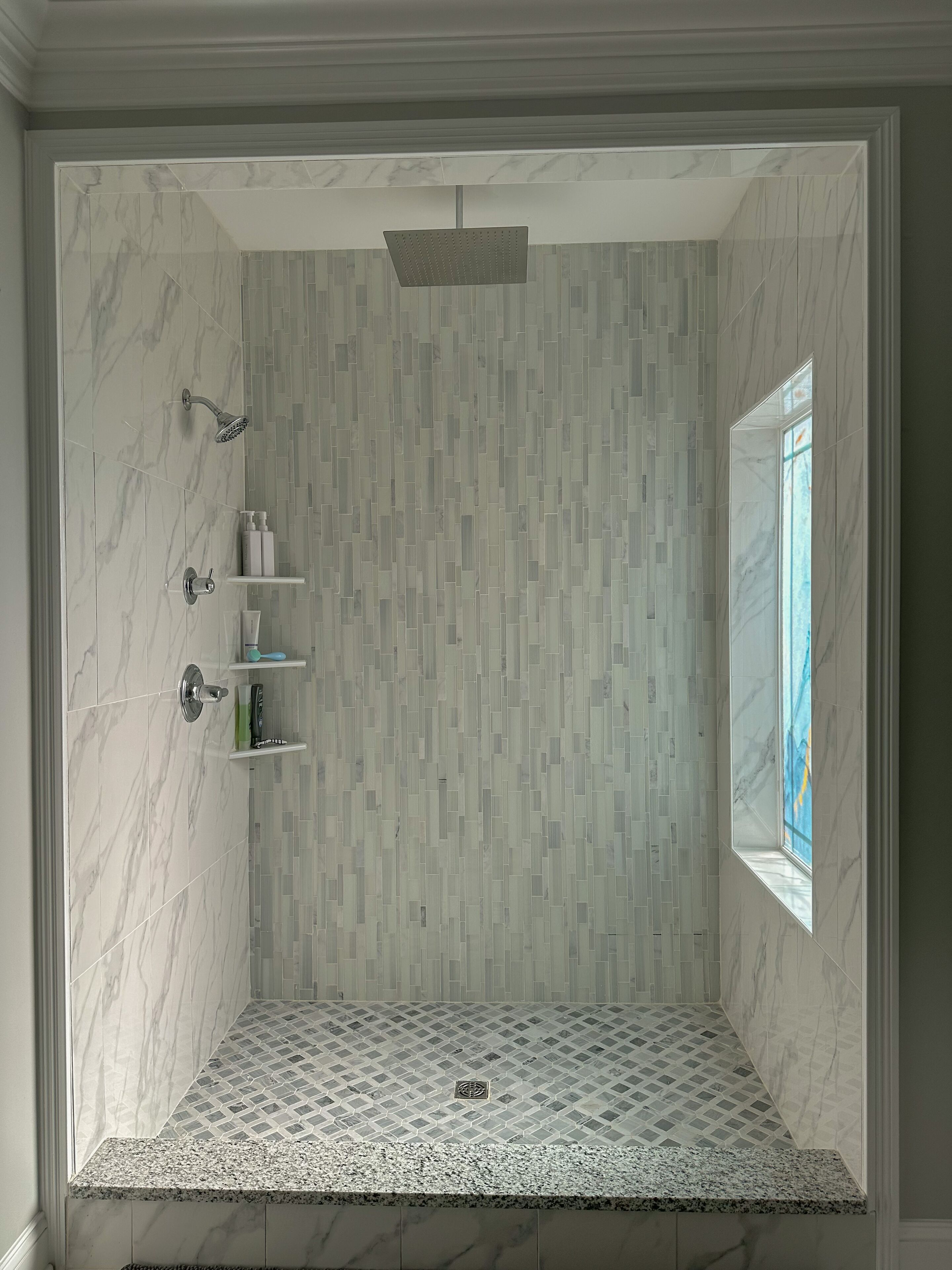 A modern shower with a rain showerhead, a small shelf with toiletries, and a window providing natural light.