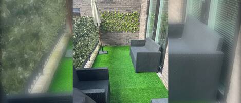 Et lille udendørs område med kunstgræs, en murstensmur og espalier med beplantning.
