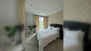 A hotel room with a bed, a potted plant, a standing fan, and a window with curtains. - Spacious & Family Friendly | 3 Beds | Stratford (London)