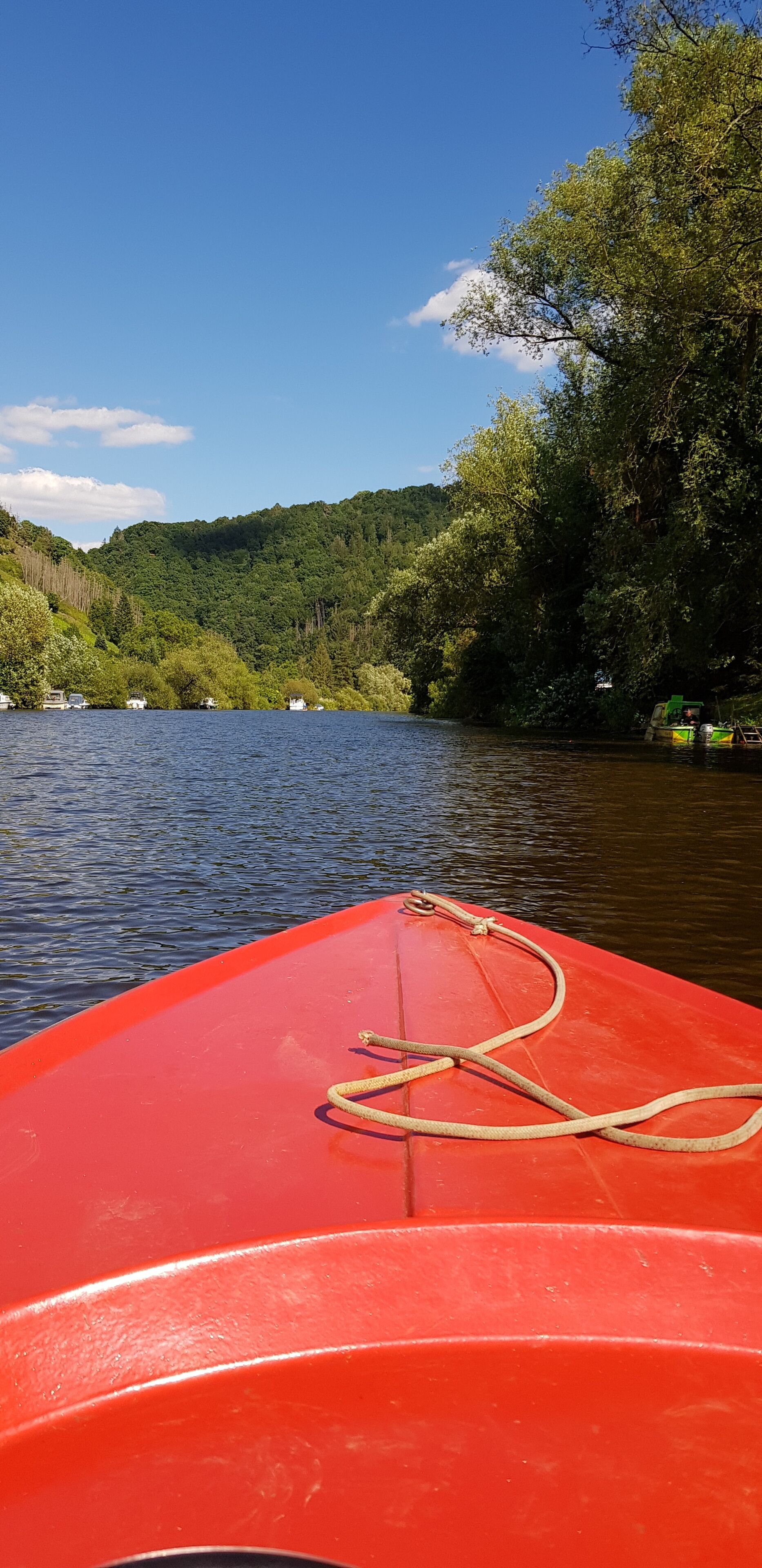 A red boat with a rope tied to its bow, floating on calm water with a forested hillside in the background.