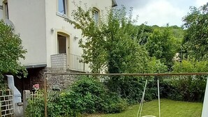 Un espace patio aménagé avec un fauteuil suspendu, une table ornée d’un vase de fleurs et une vue sur une maison à la façade en pierre et dotée d’un balcon.