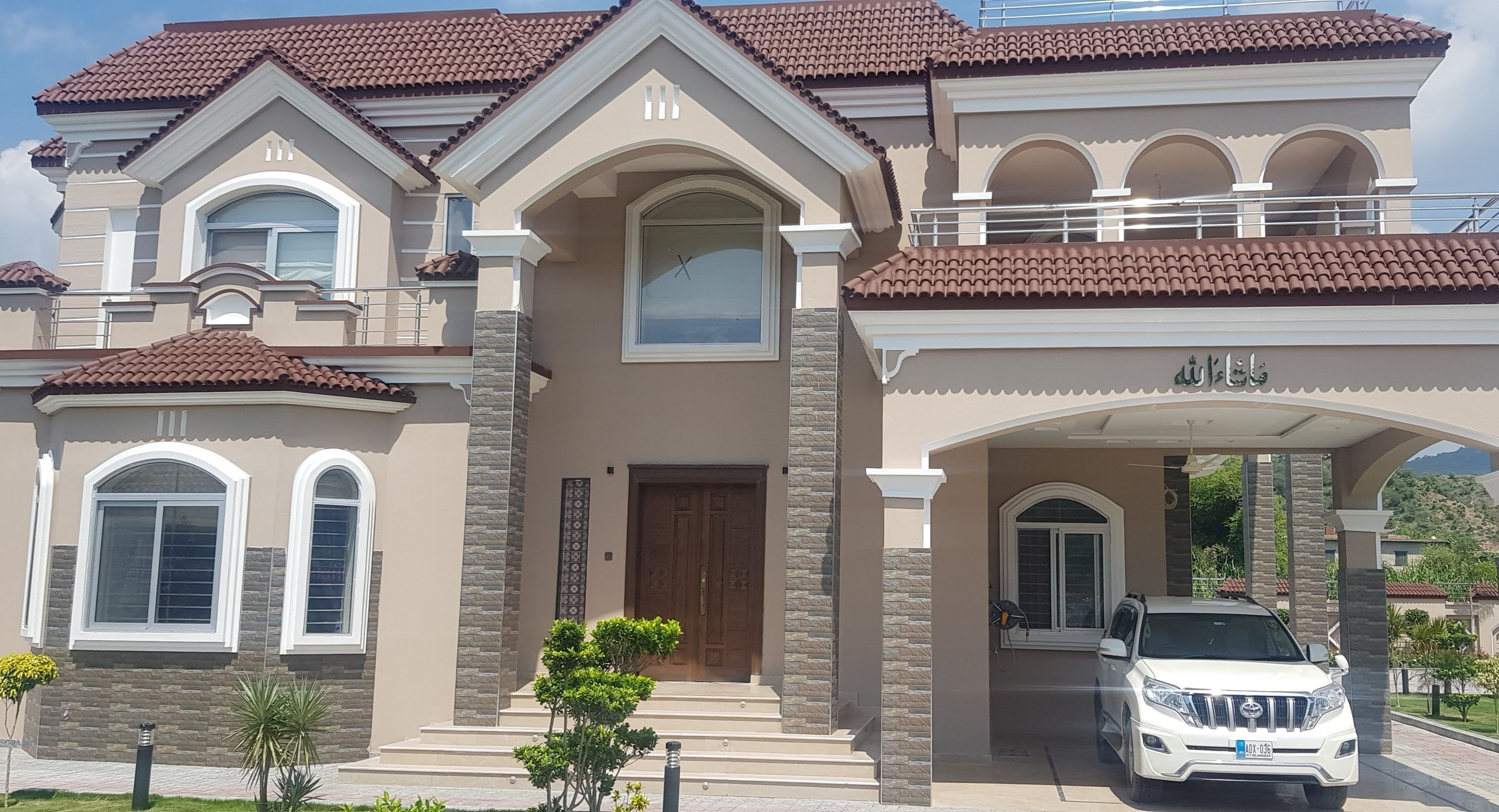 A large, two-story house with a brown tiled roof, a central entrance with a wooden door, and a white SUV parked in the driveway.