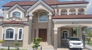 A large, two-story house with a brown tiled roof, a central entrance with a wooden door, and a white SUV parked in the driveway.