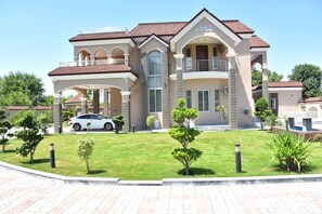 Une grande maison à deux étages, dotée d’une allée couverte, avec une voiture garée devant et une pelouse bien entretenue agrémentée de petits arbres.
