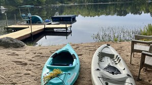 Un lago tranquilo con kayaks y una tabla de remo sobre la orilla arenosa.