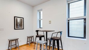 A room with a wooden floor, a table with four chairs, a framed picture on the wall, and two windows with blinds.