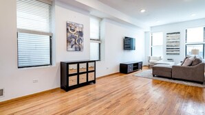 A spacious living room with hardwood flooring, a grey sofa, a coffee table, and a TV mounted on the wall.