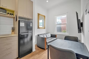 A compact kitchen with a black refrigerator, a dining table with two chairs, a grey sofa, and a window with a view of a brick building.
