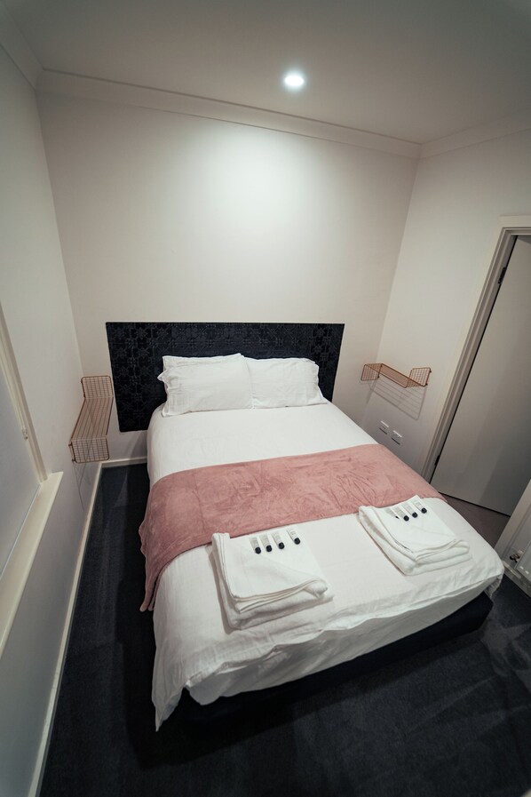 A neatly made bed with white and pink linens, a black headboard, and a bedside table with a white lamp.