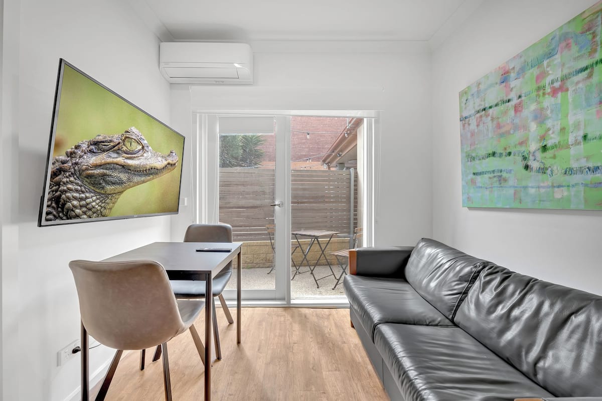 Uma sala de estar moderna com um sofá de couro, uma mesa de jantar com cadeiras, um quadro grande de um crocodilo e uma porta de vidro deslizante que dá para uma área externa.