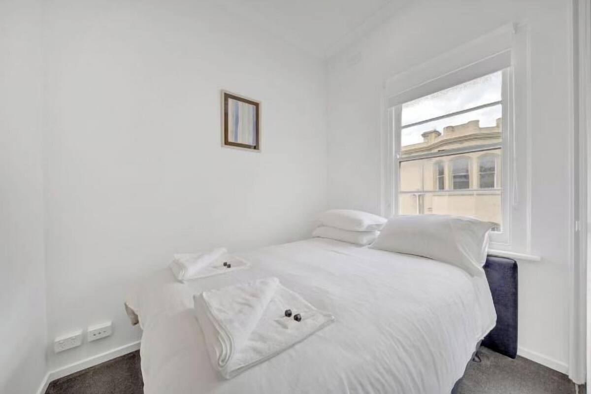 Um quarto branco com uma cama, uma janela e um pequeno quadro na parede.