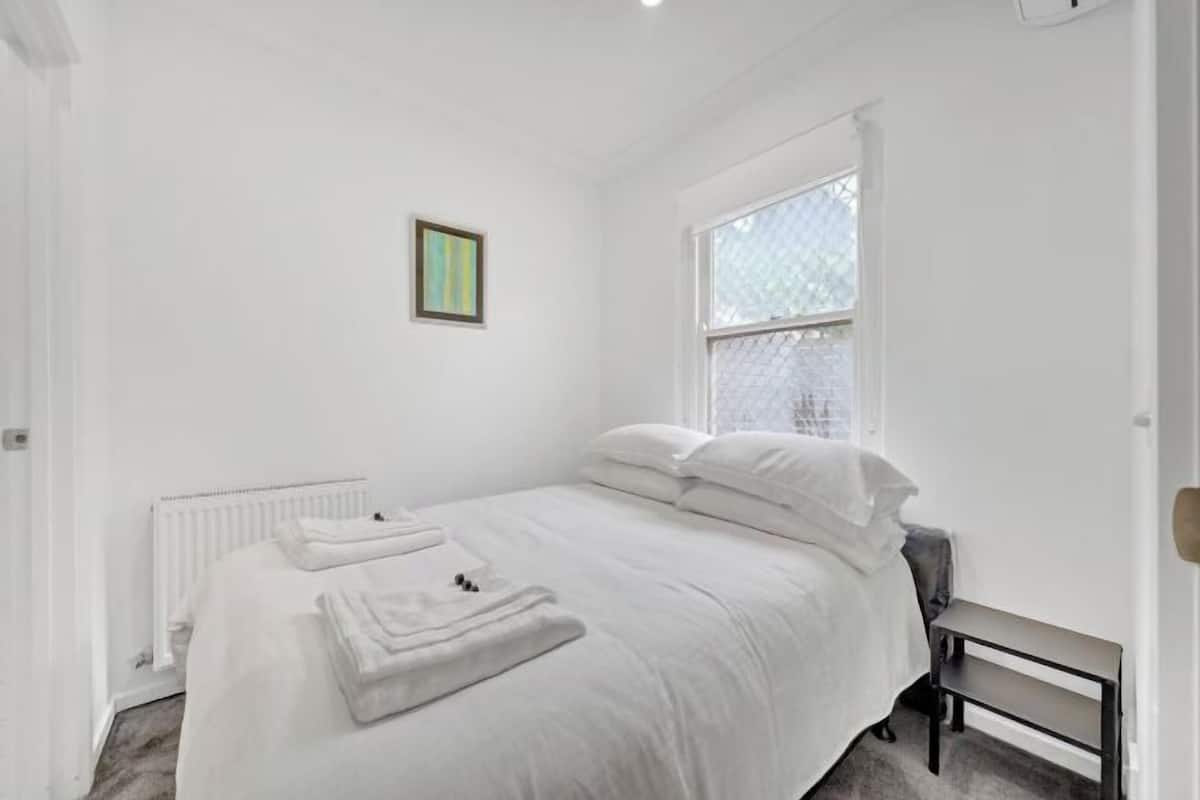 Quarto branco com cama, radiador, janela e um quadro na parede.