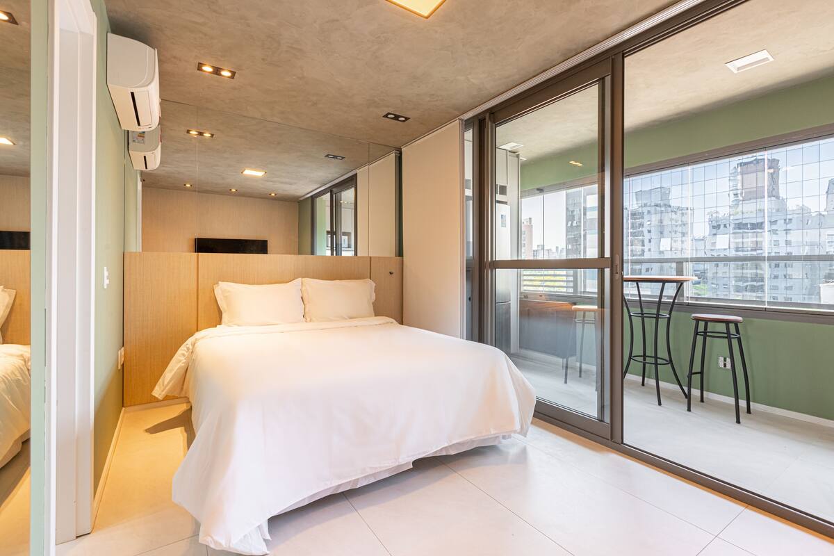 Moderni hotellihuone, jossa on suuri sänky, parvekkeella kaksi baarijakkaraa ja näkymä kaupunkiin.