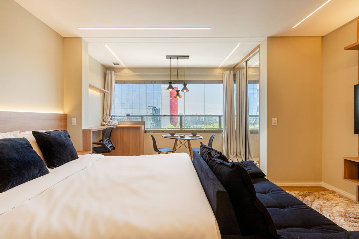Moderni hotellihuone, jossa on suuri sänky, sohva, työpöytä tuolilla sekä parveke, jolta on näkymä kaupunkiin.