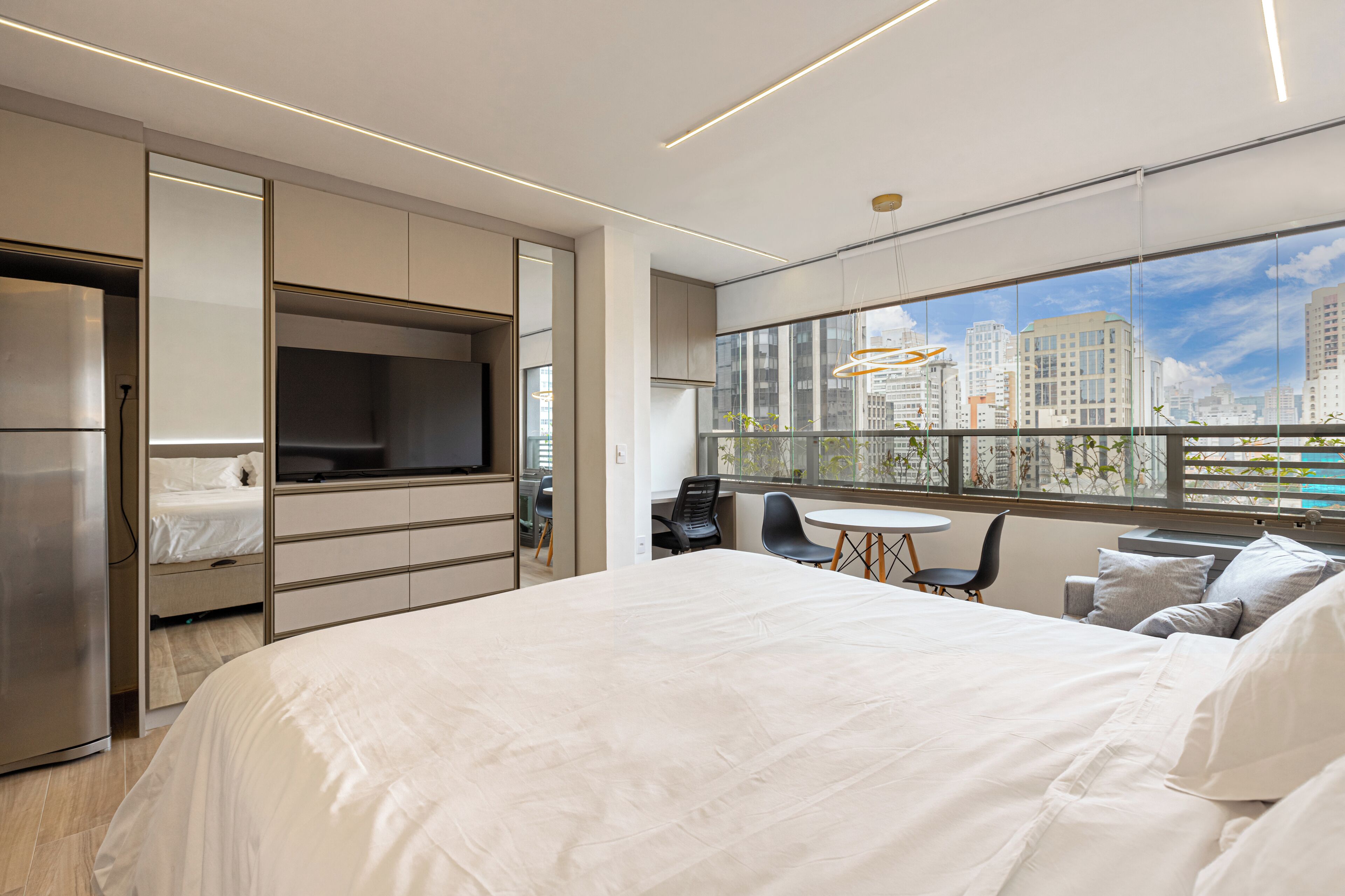 A modern hotel room with a large bed, a built-in TV, a dining area, and a city view through large windows.