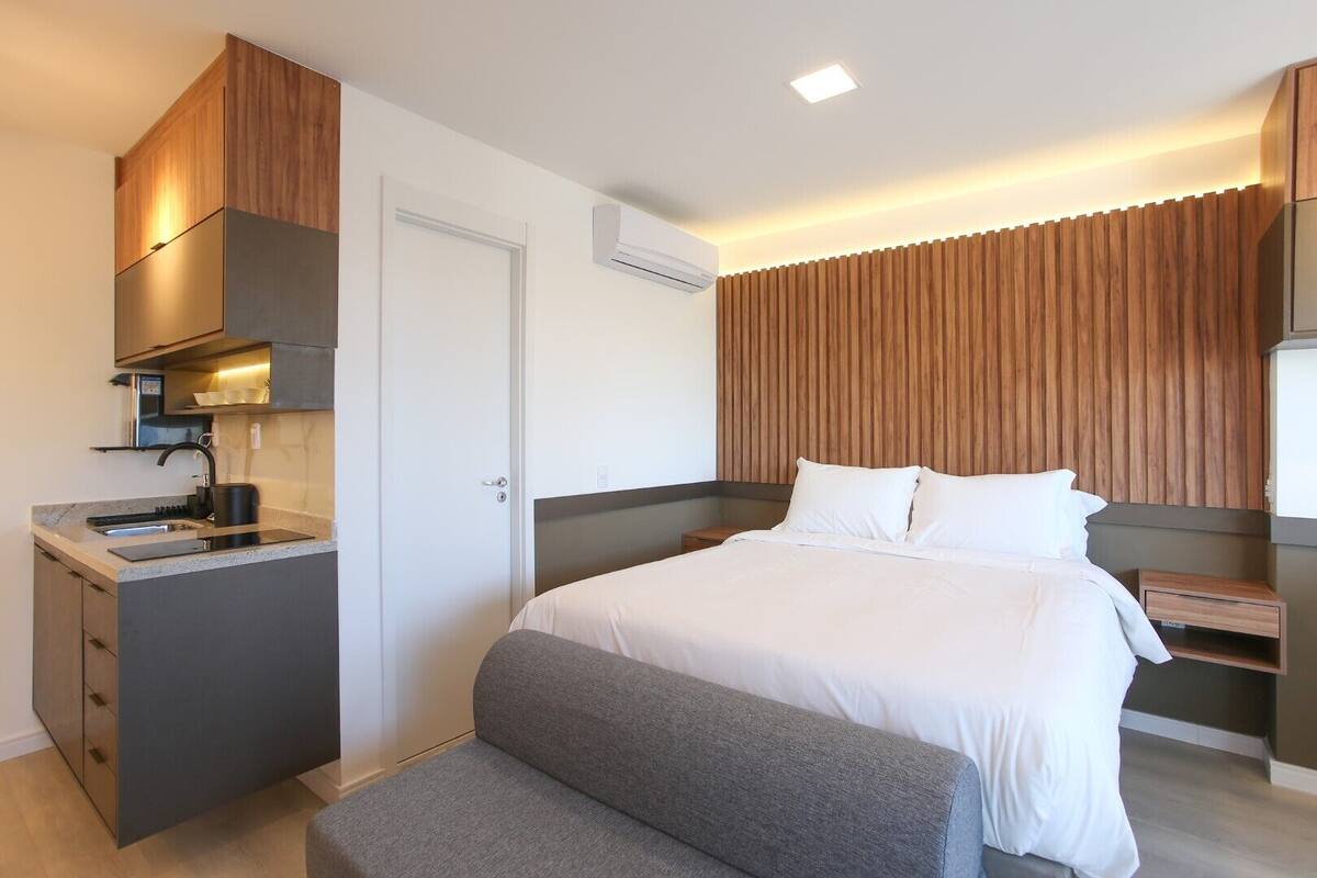 Moderni hotellihuone, jossa on pieni keittiönurkkaus, valkoisilla lakanoiden peitetty sänky, harmaa penkki ja puupanelilla verhoiltu seinä.