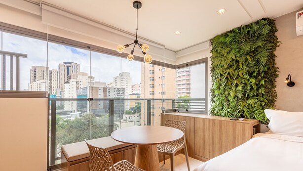 Une chambre moderne avec une grande fenêtre, une table ronde en bois, deux chaises, un lit et un mur vert orné de plantes.