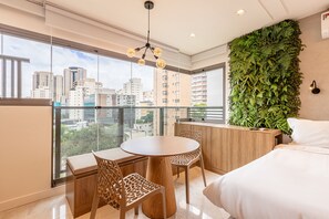 A modern bedroom with a large window, a round wooden table, two chairs, a bed, and a green wall with plants. - DSG Design Studios By Anora Spaces (São Paulo)