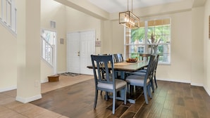 Un espace repas doté d’un plancher en bois, d’une table à manger avec des chaises, d’un lustre et d’une fenêtre avec des stores.