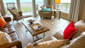 A spacious living room with a leather sofa, a brown armchair, and a beige sofa. A wooden coffee table with a tray and a decorative bird sculpture is in the center. The room has large windows with curtains, offering a view of the beach and ocean.
