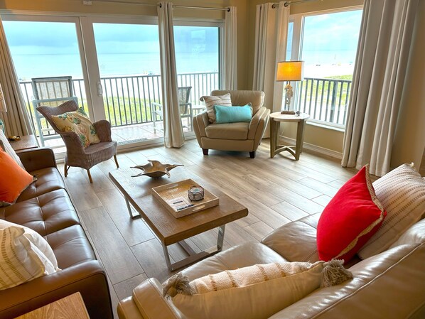 A spacious living room with a leather sofa, a brown armchair, and a beige sofa. A wooden coffee table with a tray and a decorative bird sculpture is in the center. The room has large windows with curtains, offering a view of the beach and ocean.