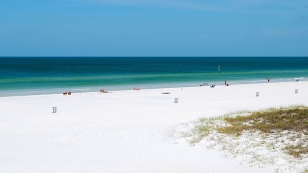 A wide, white sandy beach with clear turquoise water and a few people scattered along the shore.