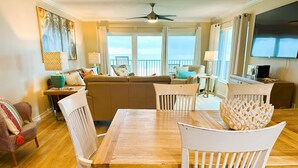 A spacious living room with a wooden dining table, white chairs, a sofa, and a view of the ocean through large windows.