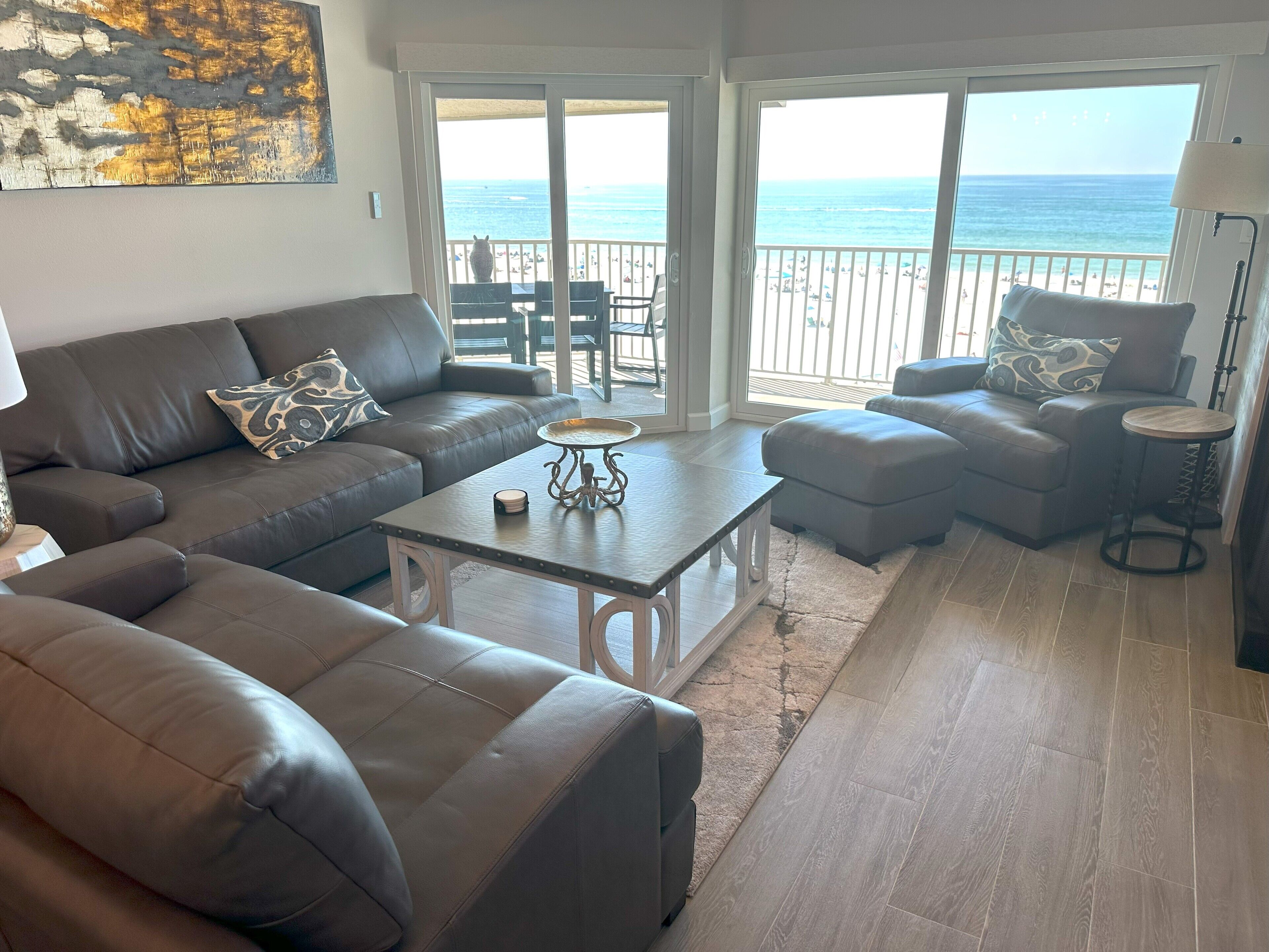 A living room with a sofa, ottoman, coffee table, and a view of the beach through large windows.