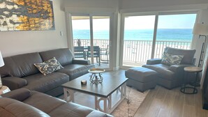Una sala de estar con sofá, reposapiés, mesa de centro y vistas a la playa a través de amplias ventanas.