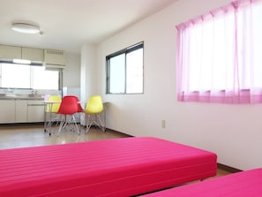 Een kamer met een kitchenette, twee stoelen, een raam met roze gordijnen en een roze bed.