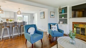 A spacious living room with a fireplace, two blue armchairs, a coffee table, and a kitchen area with white cabinets and bar stools. - Families Welcome! Home 2 Mi to Craigville Beach (Hyannis)