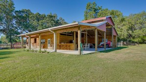 A spacious wooden building with an open front and covered patio area, surrounded by a grassy field and trees. - Farm Stay: Modern Barndominium in Lakeland! (Lakeland)
