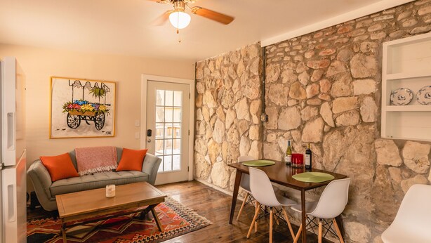 A cozy living room with a stone wall, a wooden dining table with chairs, a sofa with orange cushions, and a ceiling fan.