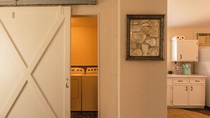A laundry room with a washing machine and dryer, a framed artwork on the wall, and a kitchenette area with cabinets and a coffee maker.