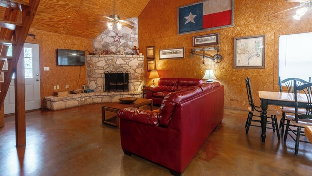 A spacious living room with a stone fireplace, a red leather couch, a dining table with chairs, and a Texas flag on the wall. - Located 1/4 Mile to River Access (Concan)