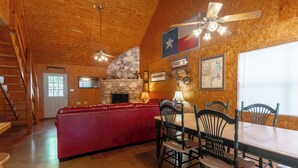 A cozy cabin interior with a stone fireplace, wooden ceiling, a red couch, and a dining area with wooden chairs and a table. - Located 1/4 Mile to River Access (Concan)