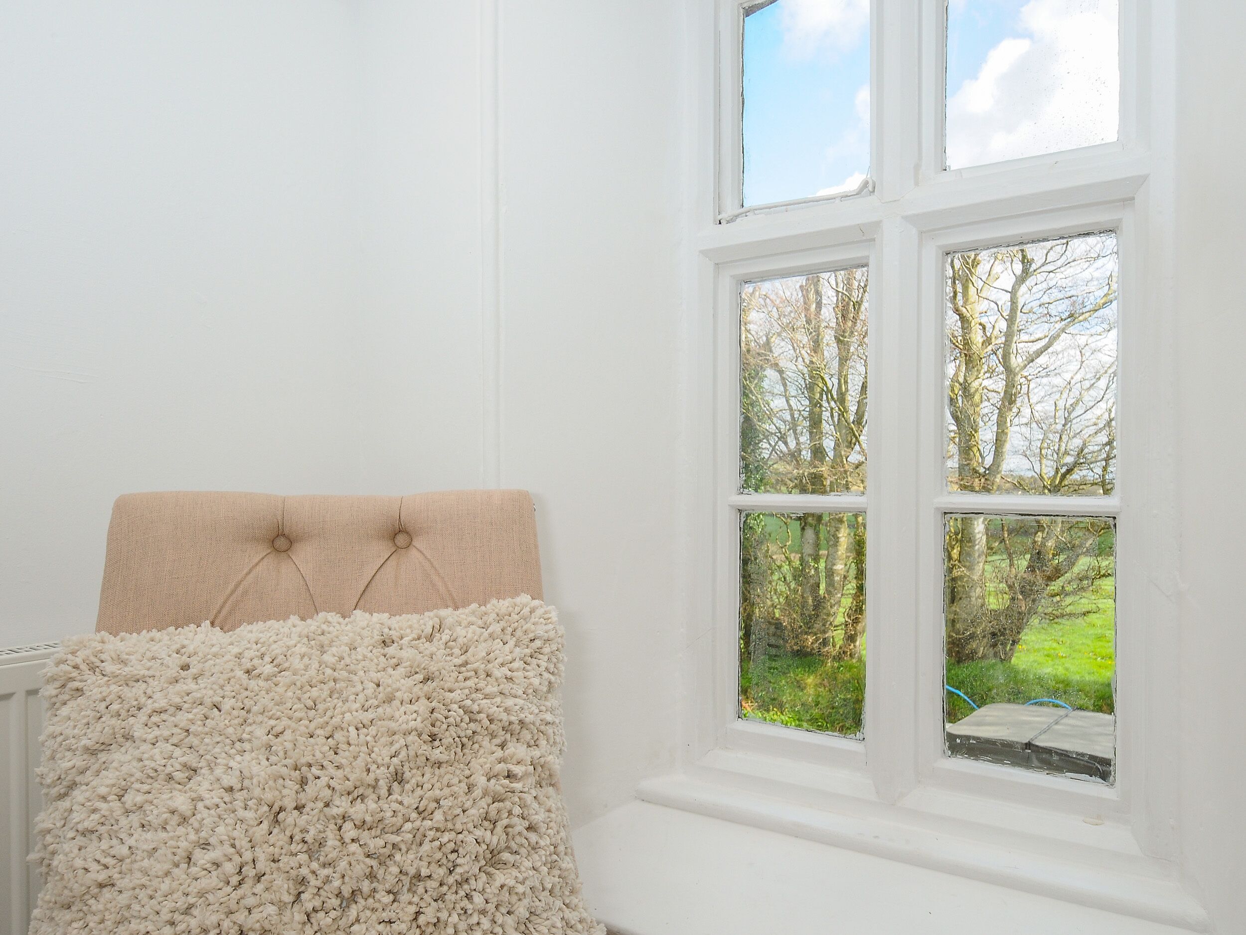 A room with a window, a white wall, and a beige headboard with a fluffy white throw.