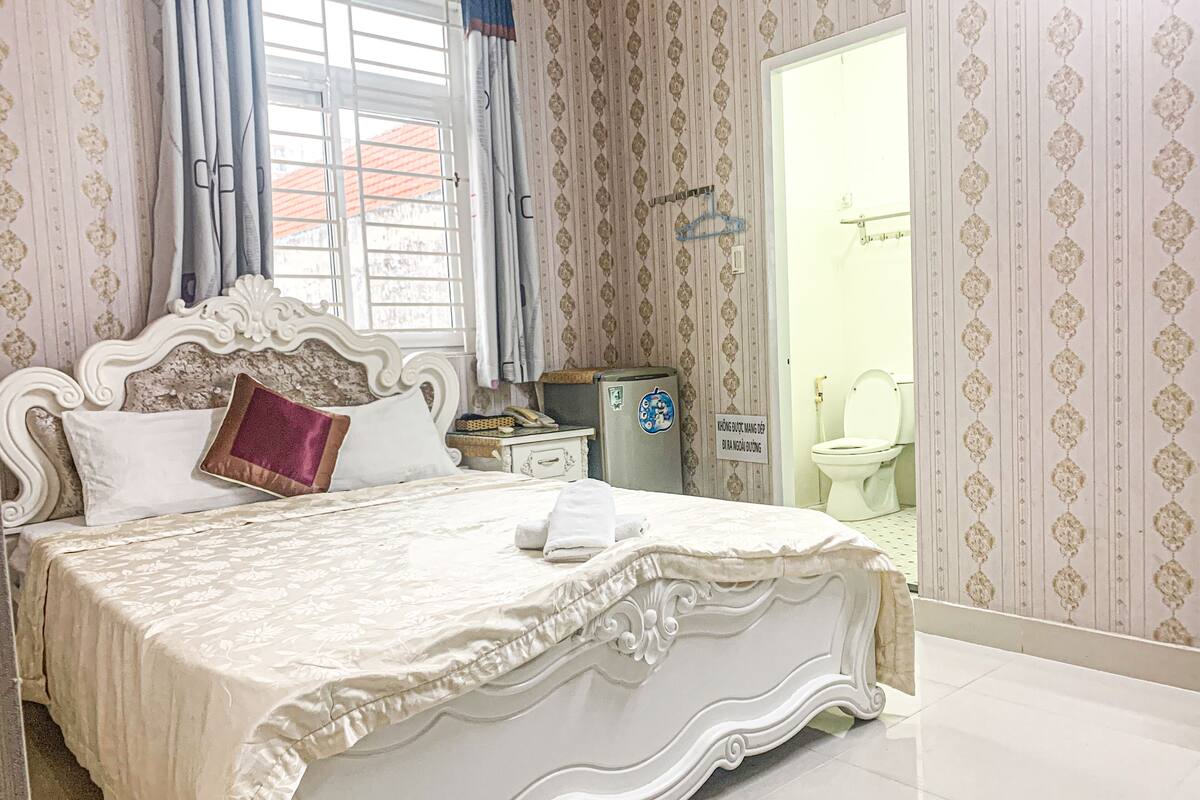 Une chambre à coucher avec un grand lit, une table de chevet, un réfrigérateur et une salle de bain visible à travers une porte ouverte.