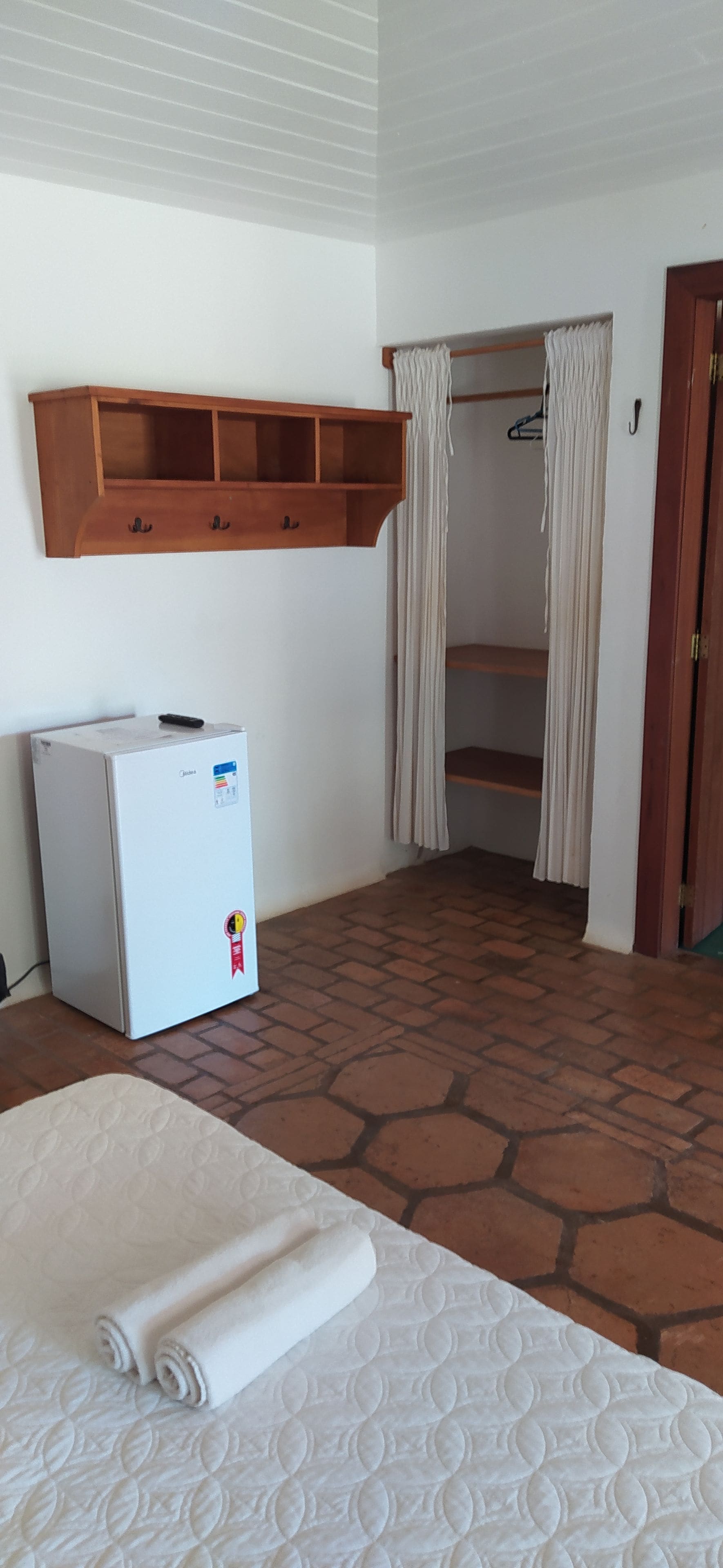 A room with a bed, a small refrigerator, a wooden shelf, and a closet with hanging clothes.