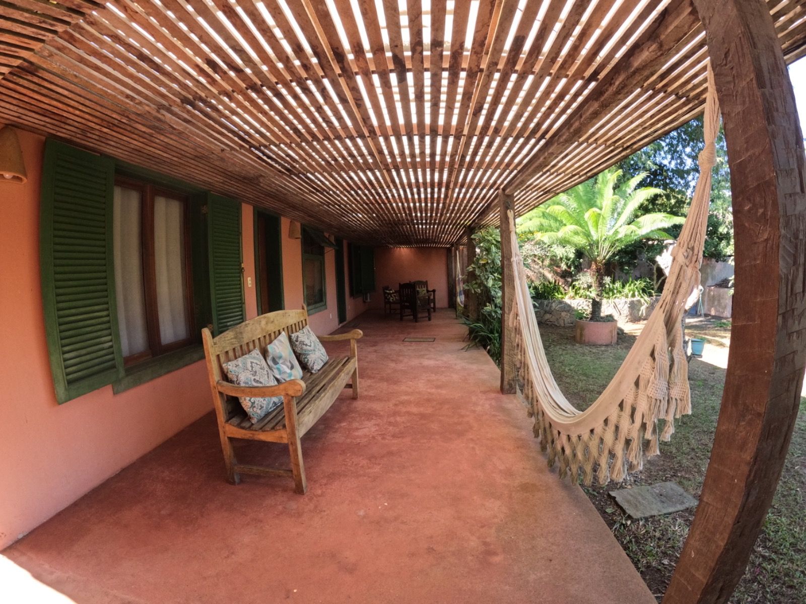 A covered outdoor area with a wooden bench, a hammock, and a view of a garden with palm trees.