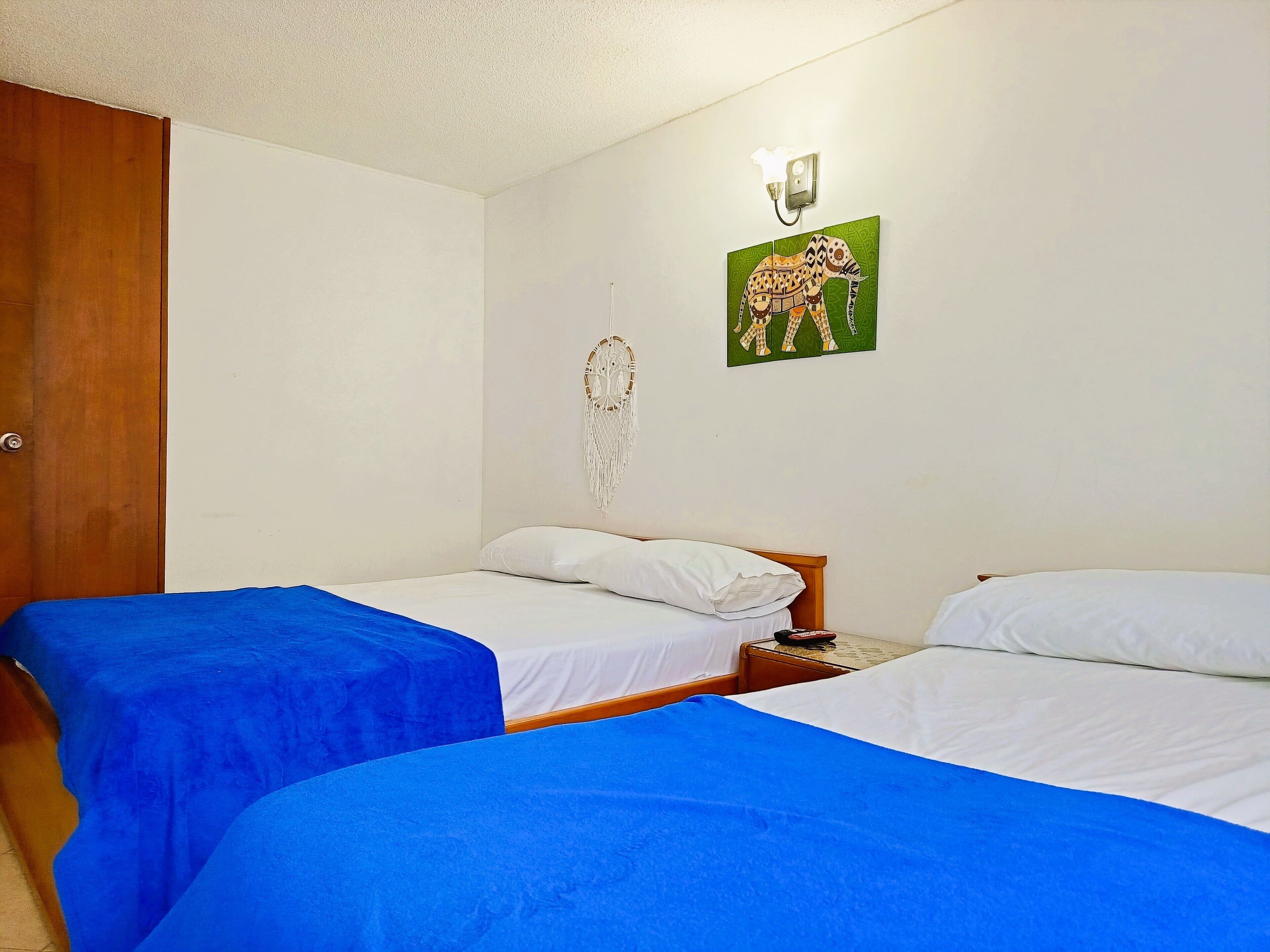 A room with two beds, each with a blue bedspread and white bedding. There is a wooden door on the left, a wall-mounted light fixture, and a wall decoration of an elephant.