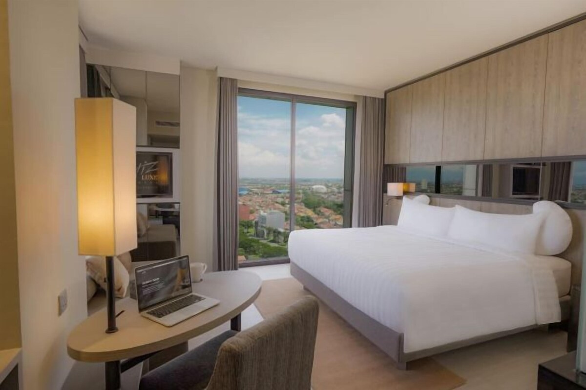 A hotel room with a large bed, a desk with a laptop, a chair, and a view of the city through large windows.