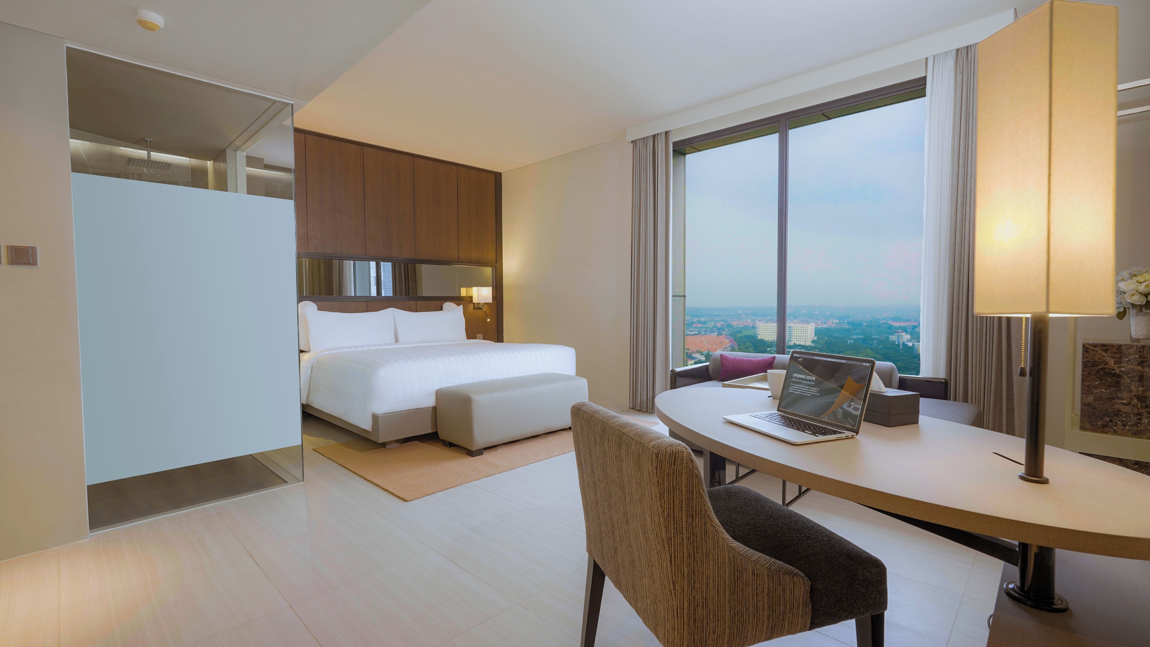 Quarto de hotel com cama, secretária com um laptop, cadeira e vista da cidade através de grandes janelas.