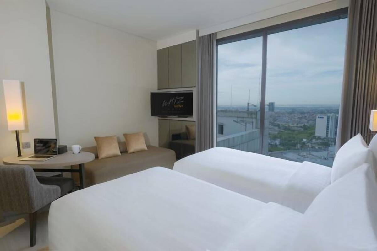 Une chambre d’hôtel avec deux lits, une télévision, un bureau avec une chaise, un canapé et une grande fenêtre donnant sur la ville.