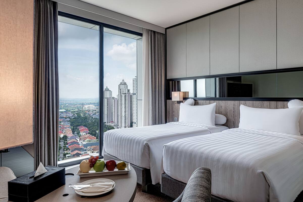 Une chambre d’hôtel avec deux lits, une grande fenêtre donnant sur la ville, une table de chevet avec un plateau de fruits et un téléviseur fixé au mur.